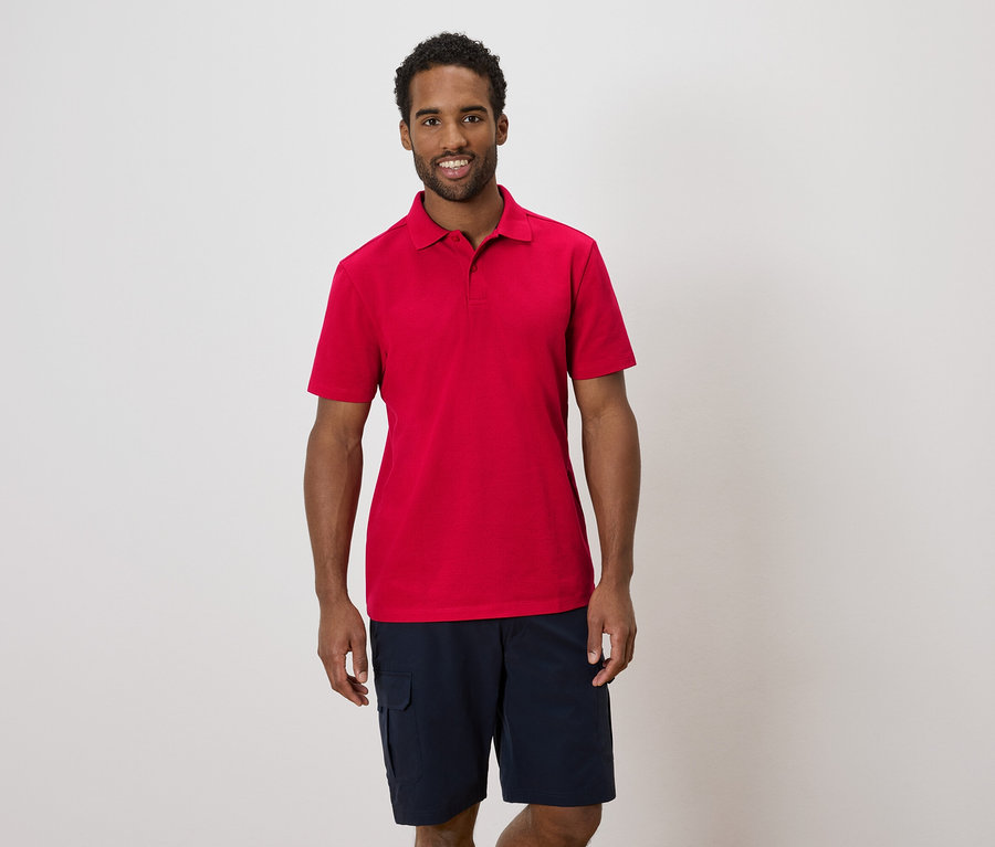 Un homme portant un polo rouge et un short bleu se tient devant un mur blanc.