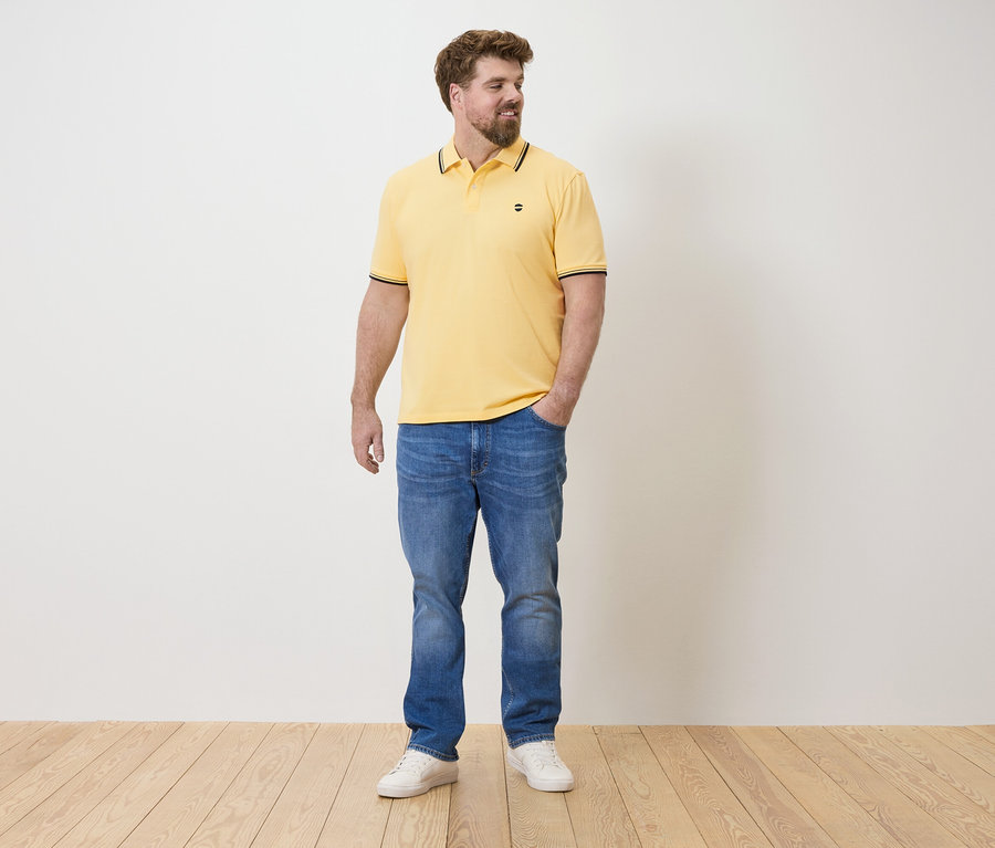 Un homme se tient debout, une main dans la poche, portant un polo jaune, un jean bleu et des baskets blanches.