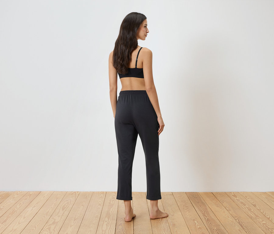 Femme debout dos à la caméra, portant un soutien-gorge noir et un pantalon foncé.