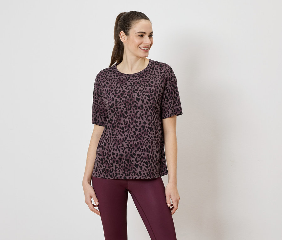 Une femme porte un t-shirt violet à motif léopard et un legging violet sur un fond blanc.