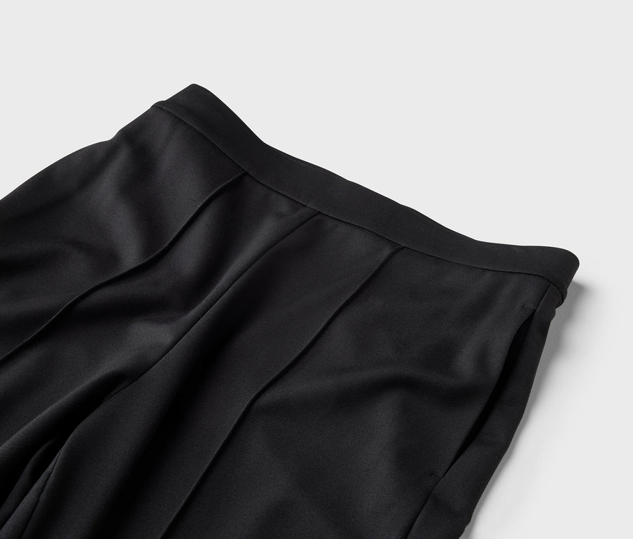 Gros plan sur la taille d'un pantalon noir avec des plis et une poche latérale sur fond gris.