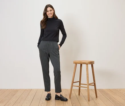 Une femme portant un pull à col roulé noir et un pantalon rayé pose à côté d'un tabouret sur un plancher en bois.