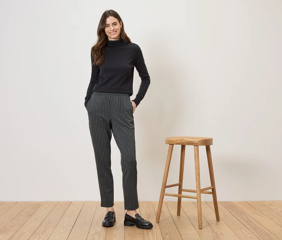 Une femme portant un pull à col roulé noir et un pantalon rayé pose à côté d'un tabouret sur un plancher en bois.