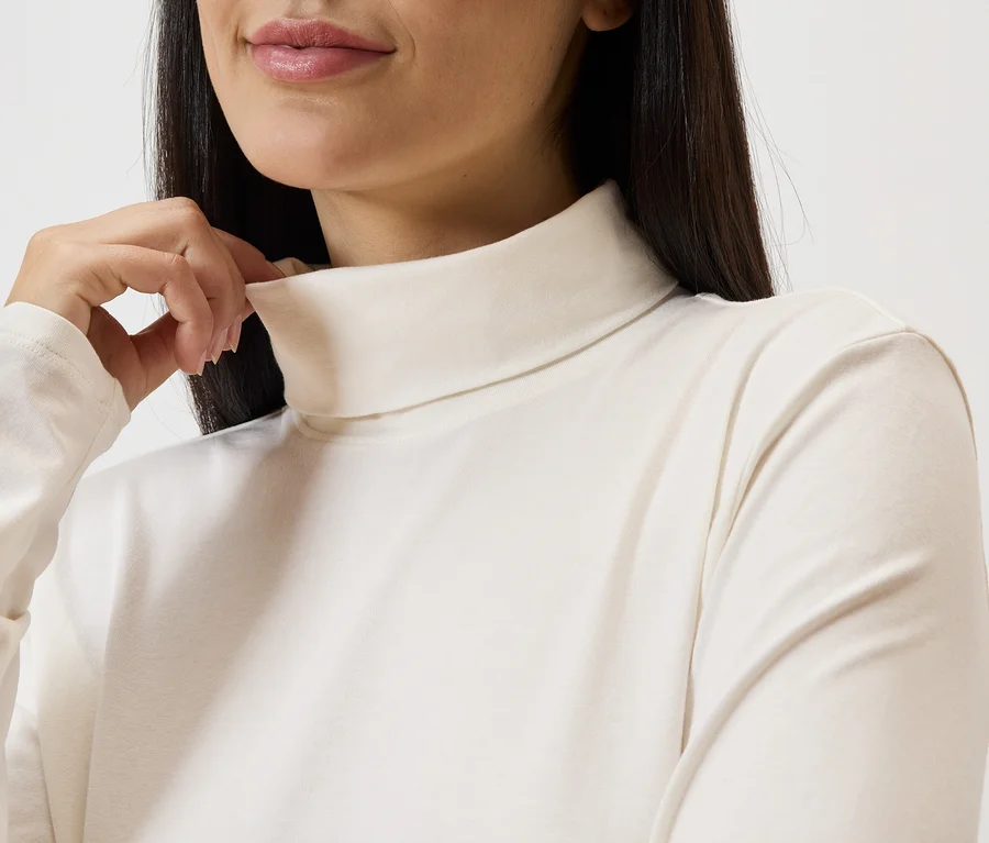 Une femme touche son col roulé. Le haut est à manches longues et de couleur claire.