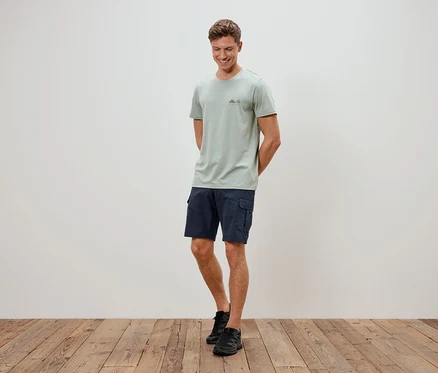 Un homme debout devant un mur portant un t-shirt vert clair, un short bleu et des chaussures noires.