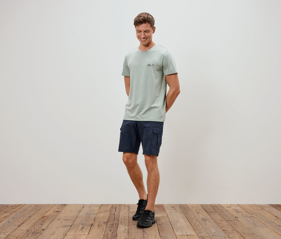 Un homme debout devant un mur portant un t-shirt vert clair, un short bleu et des chaussures noires.