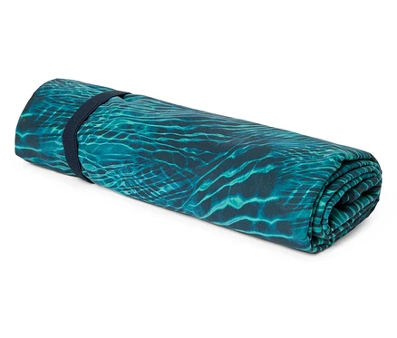 Un tapis de plage turquoise enroulé avec un motif qui rappelle les ondulations de l'eau, maintenu par un élastique bleu marine.
