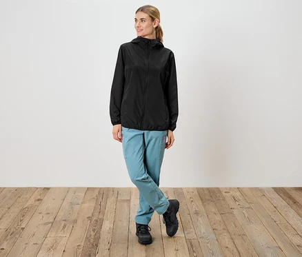 Une femme portant un imperméable noir à capuche, un pantalon bleu clair et des chaussures noires se tient devant un mur blanc.