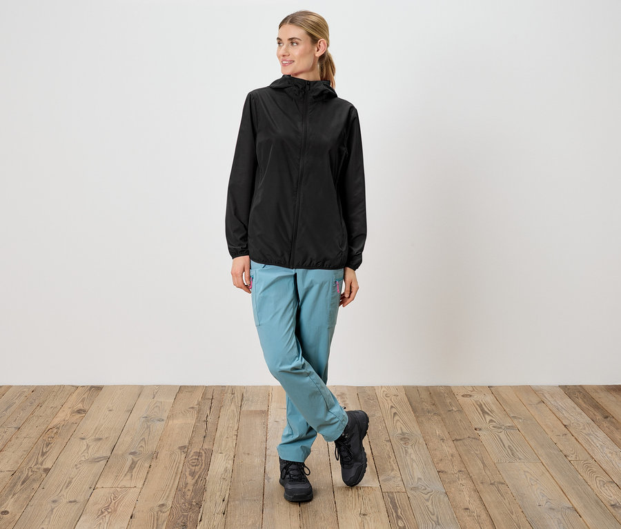 Une femme portant un imperméable noir à capuche, un pantalon bleu clair et des chaussures noires se tient devant un mur blanc.