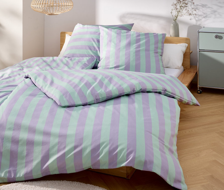 Un lit avec des draps rayés violets et verts se trouve dans une pièce lumineuse.