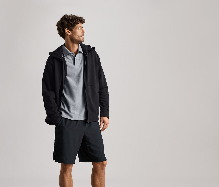 Un homme porte une veste de survêtement noire sur un polo gris et un short noir.