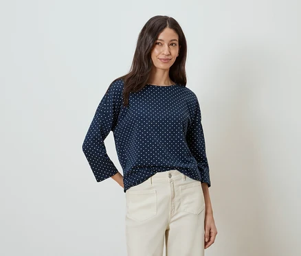 Une femme aux longs cheveux bruns porte un chemisier bleu marine à pois blancs et un pantalon beige.
