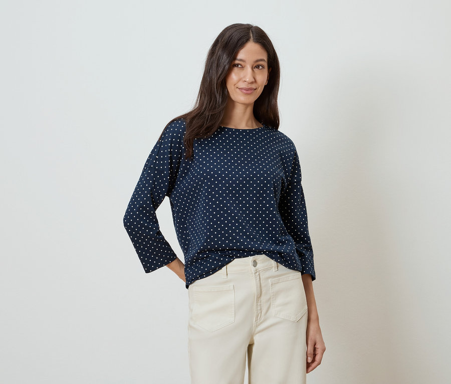 Une femme aux longs cheveux bruns porte un chemisier bleu marine à pois blancs et un pantalon beige.