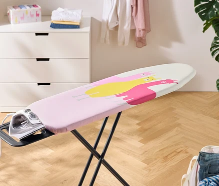 Un fer à repasser repose sur une table à repasser recouverte d'une housse rose à motifs de lamas jaunes et roses. À l'arrière-plan, une commode, du linge et une plante d'intérieur.