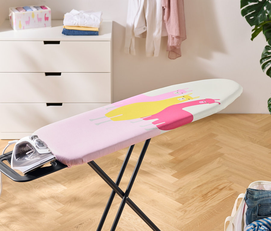 Un fer à repasser repose sur une table à repasser recouverte d'une housse rose à motifs de lamas jaunes et roses. À l'arrière-plan, une commode, du linge et une plante d'intérieur.