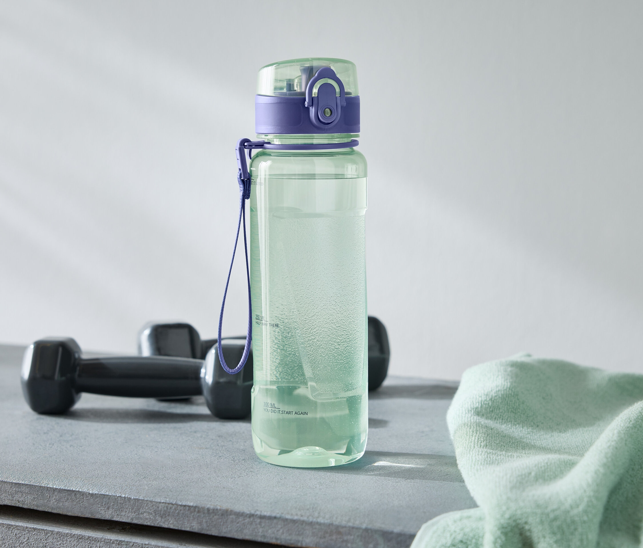 Une bouteille d'eau verte avec un bouchon violet se trouve à côté de deux haltères noirs et d'une serviette vert clair sur une surface grise.