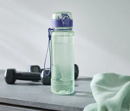 Une bouteille d'eau verte avec un bouchon violet se trouve à côté de deux haltères noirs et d'une serviette vert clair sur une surface grise.