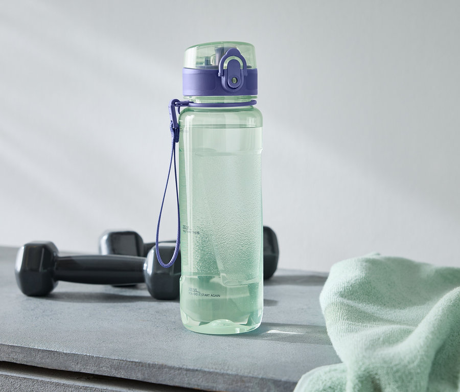 Une bouteille d'eau verte avec un bouchon violet se trouve à côté de deux haltères noirs et d'une serviette vert clair sur une surface grise.