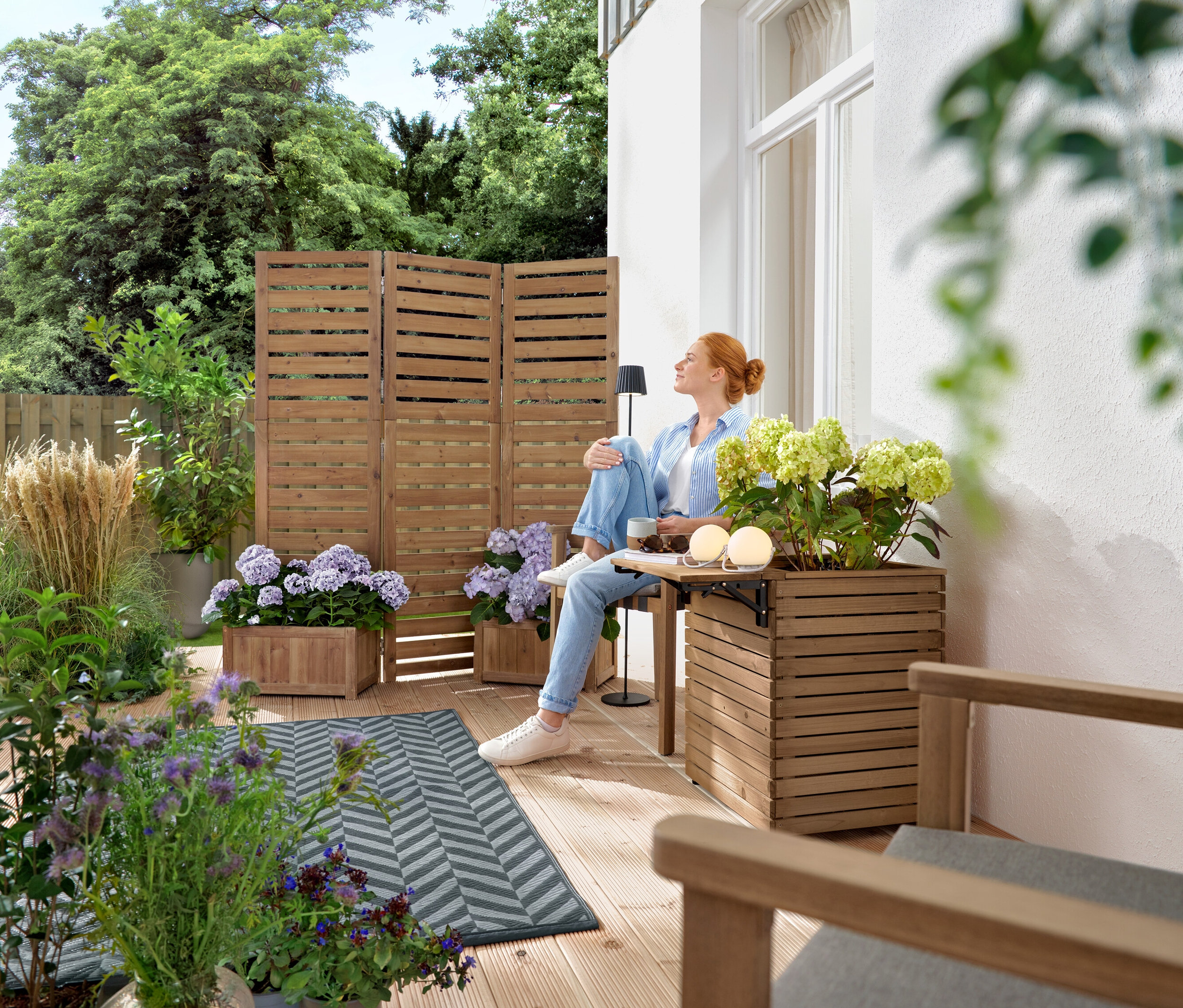 Femme assise sur une terrasse en bois avec table et boisson, entourée de plantes dans des caisses en bois.