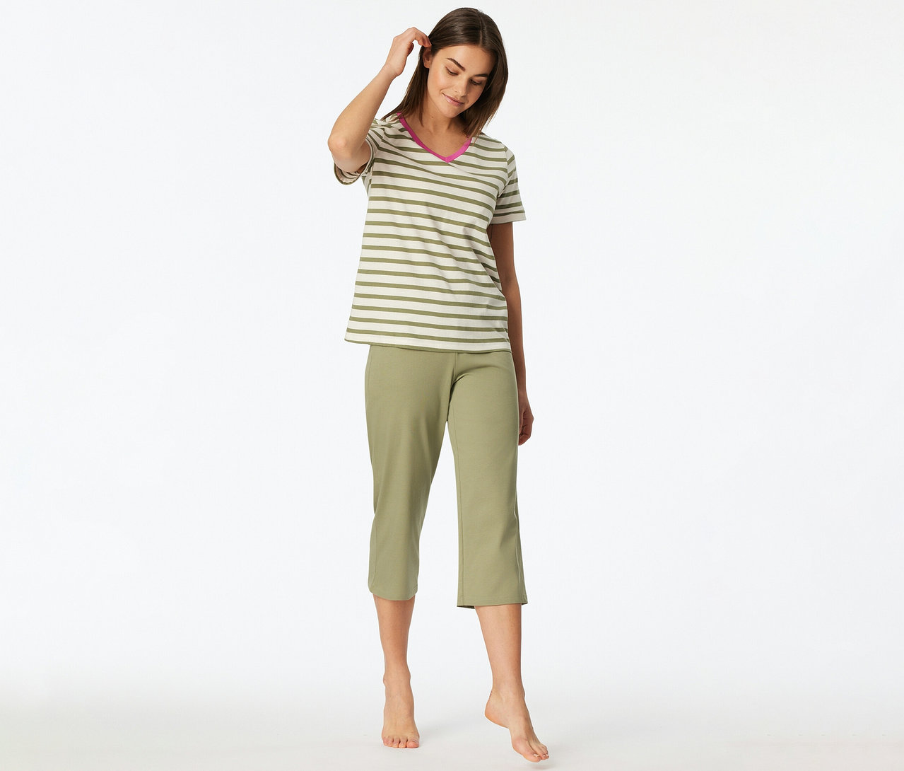 Une femme est debout pieds nus, regardant vers le bas et touchant ses cheveux, vêtue d'un Set de pyjama 3/4 SCHIESSER composé d'un haut rayé à manches courtes avec un col en V rose et d'un pantalon 3/4 vert olive.