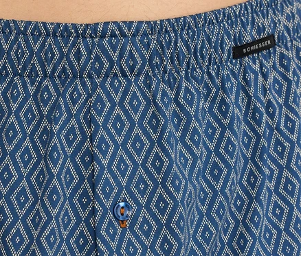 Gros plan sur un boxer en jersey SCHIESSER bleu avec un motif de losanges blancs, une ceinture élastique, une étiquette SCHIESSER noire et un bouton bleu avec des coutures oranges.