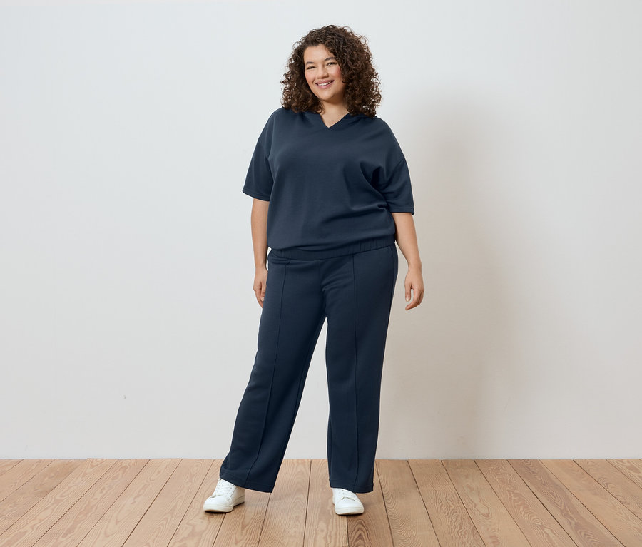 Femme aux cheveux bouclés portant un t-shirt et un pantalon bleu marine devant un mur blanc.