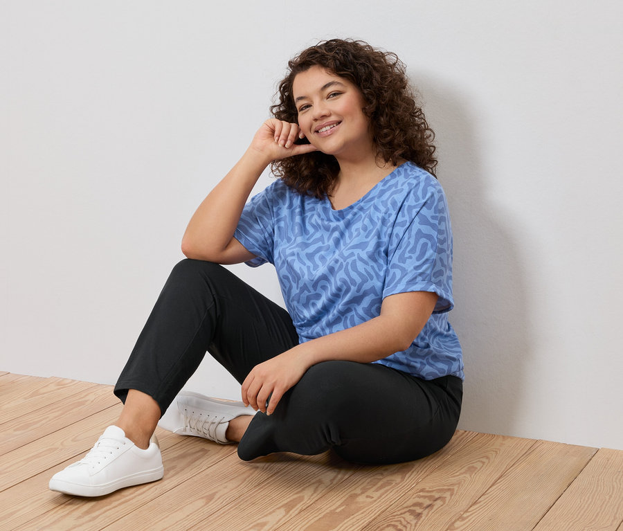 Femme aux cheveux bouclés assise sur un plancher en bois, portant un t-shirt bleu à motifs, un pantalon noir et des baskets blanches.