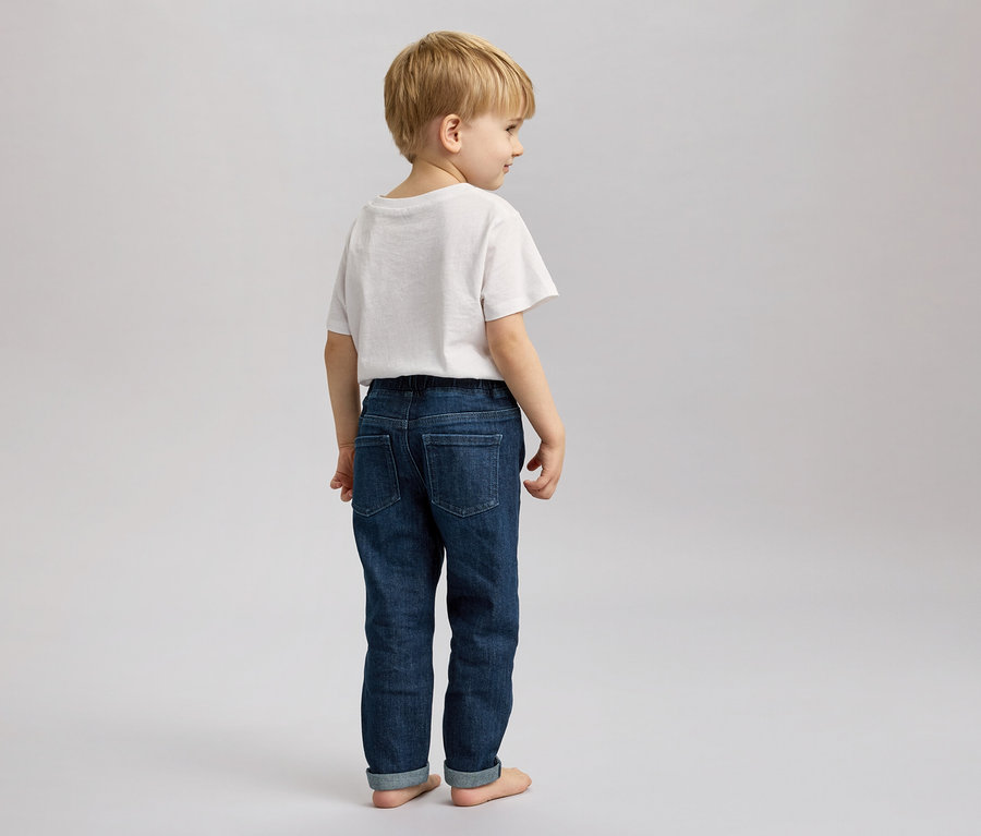 Un garçon blond en t-shirt blanc et jean est pieds nus sur un fond gris et regarde à droite.