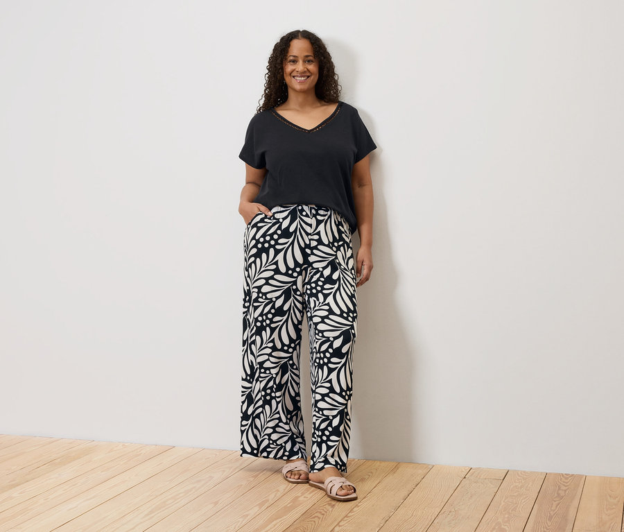 Une femme souriante aux cheveux bouclés, portant un t-shirt noir et un pantalon à motifs, se tient devant un mur blanc.