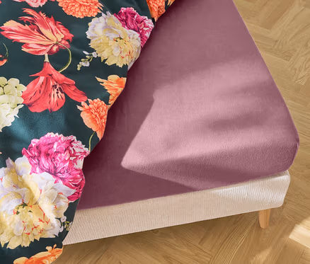 Gros plan d'un lit avec un drap-housse violet et une couette à fleurs sur un plancher en bois.