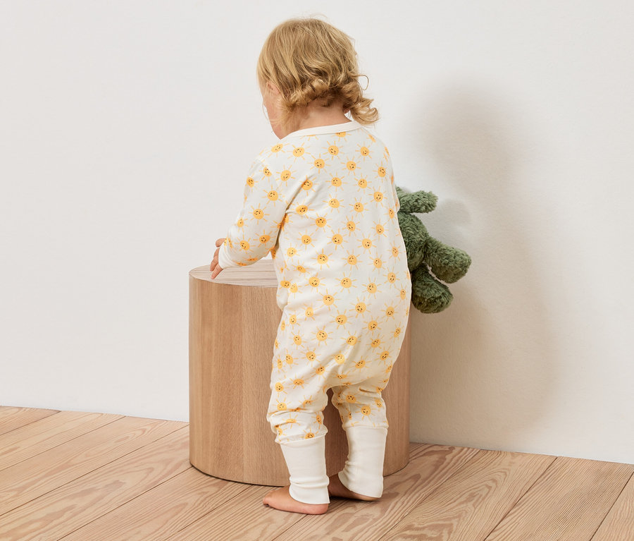 Un jeune enfant en combinaison jaune et blanche à motifs de soleil se tient à côté d'un tabouret rond en bois, avec une peluche verte à côté.