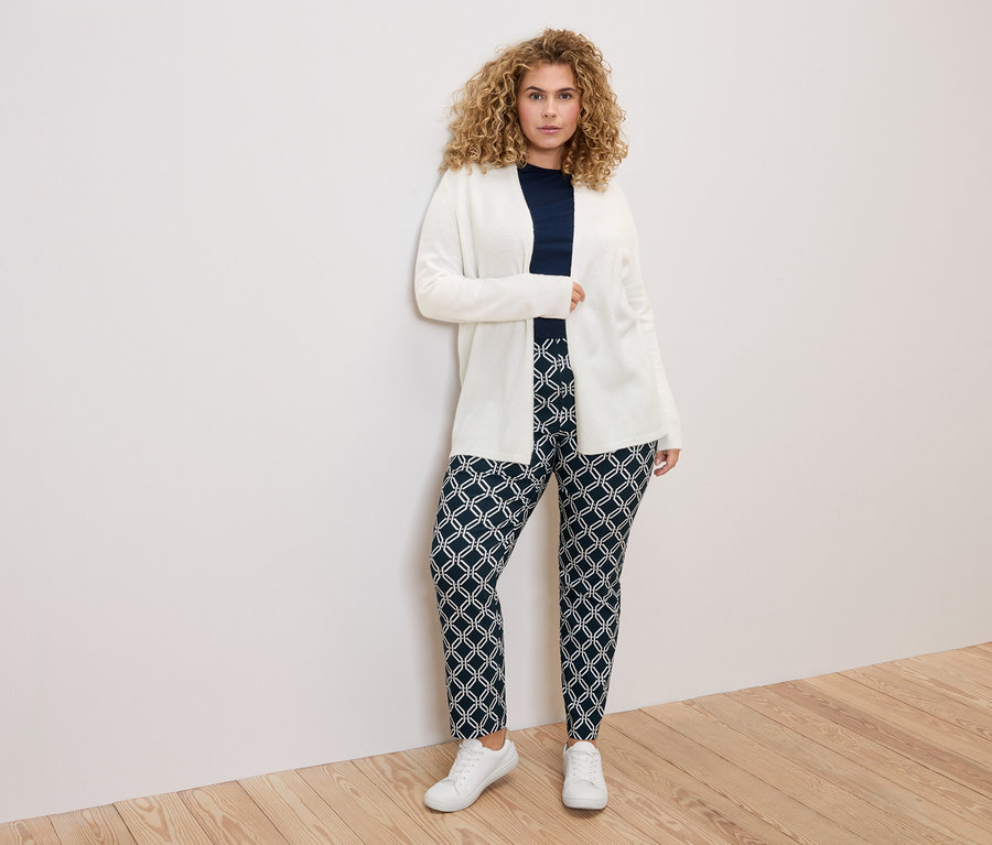 Une femme aux cheveux bouclés portant un cardigan blanc, un haut bleu marine et un pantalon à motifs devant un mur blanc.