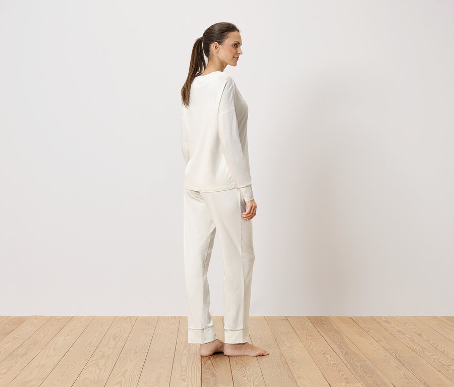 Femme debout de profil en pyjama crème à manches longues devant un mur blanc.