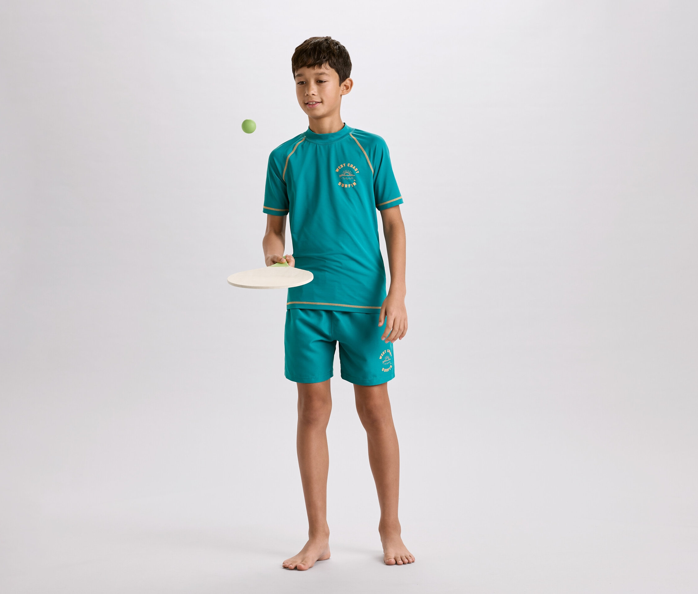 Un jeune garçon pieds nus, vêtu d'un t-shirt turquoise et d'un short avec des accents marron clair, joue avec une balle de paddle sur un fond gris clair.