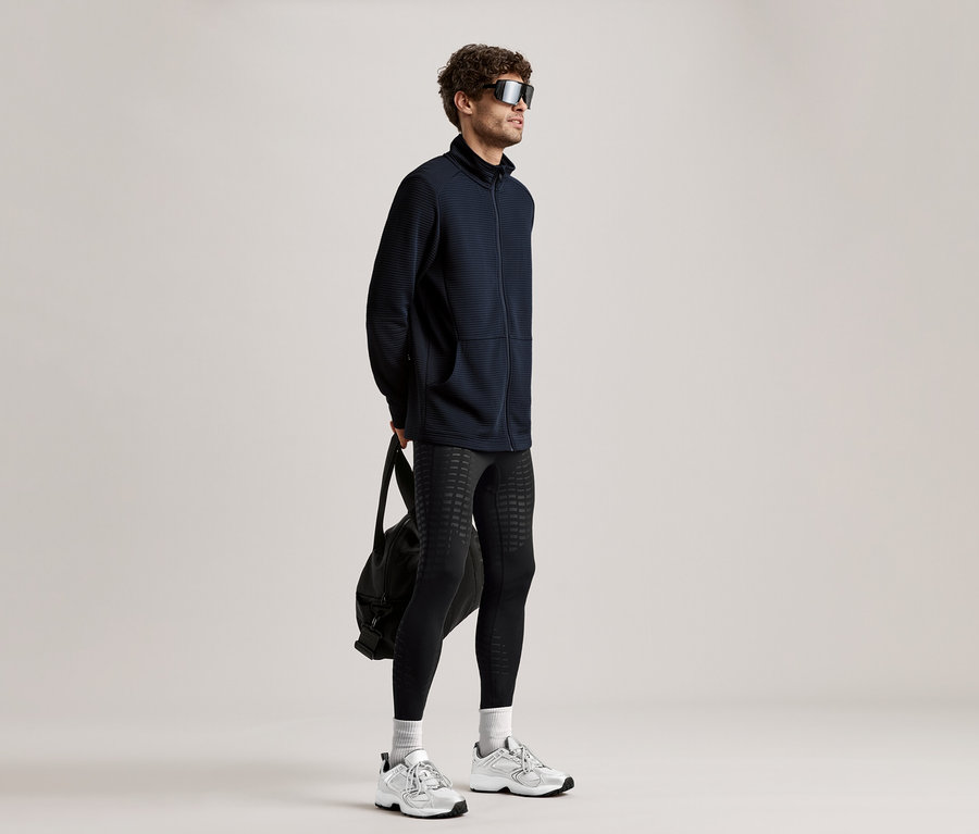 Un homme portant des lunettes de soleil, une veste noire, des leggings et des baskets tient un sac noir.