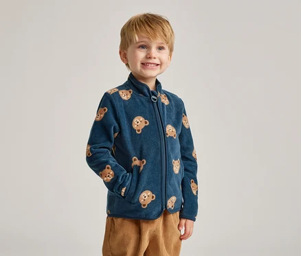 Un garçon souriant aux cheveux blonds porte une veste en polaire bleu marine à motifs d'ours et un pantalon en velours côtelé marron.