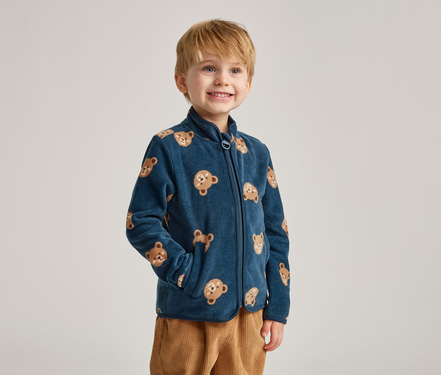 Un garçon souriant aux cheveux blonds porte une veste en polaire bleu marine à motifs d'ours et un pantalon en velours côtelé marron.