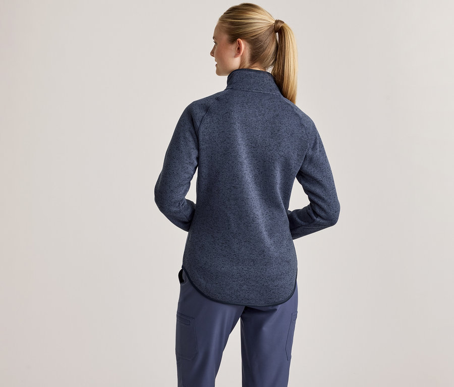 Une femme vue de dos, les cheveux blonds attachés en queue de cheval, porte une veste polaire mouchetée bleue à col montant et un pantalon bleu.