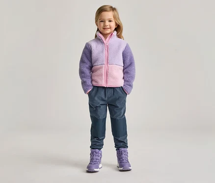 Une fillette portant une veste en polaire violette et rose, un pantalon bleu et des bottes violettes se tient devant un fond clair.