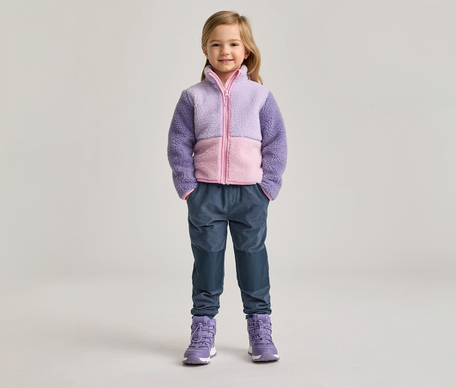 Une fillette portant une veste en polaire violette et rose, un pantalon bleu et des bottes violettes se tient devant un fond clair.
