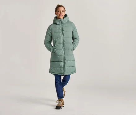 Une femme en jean et bottes porte un long manteau matelassé vert menthe à capuche, les mains dans les poches.