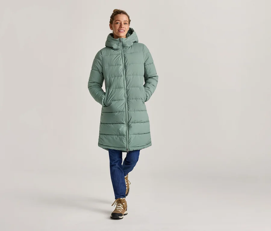Une femme en jean et bottes porte un long manteau matelassé vert menthe à capuche, les mains dans les poches.