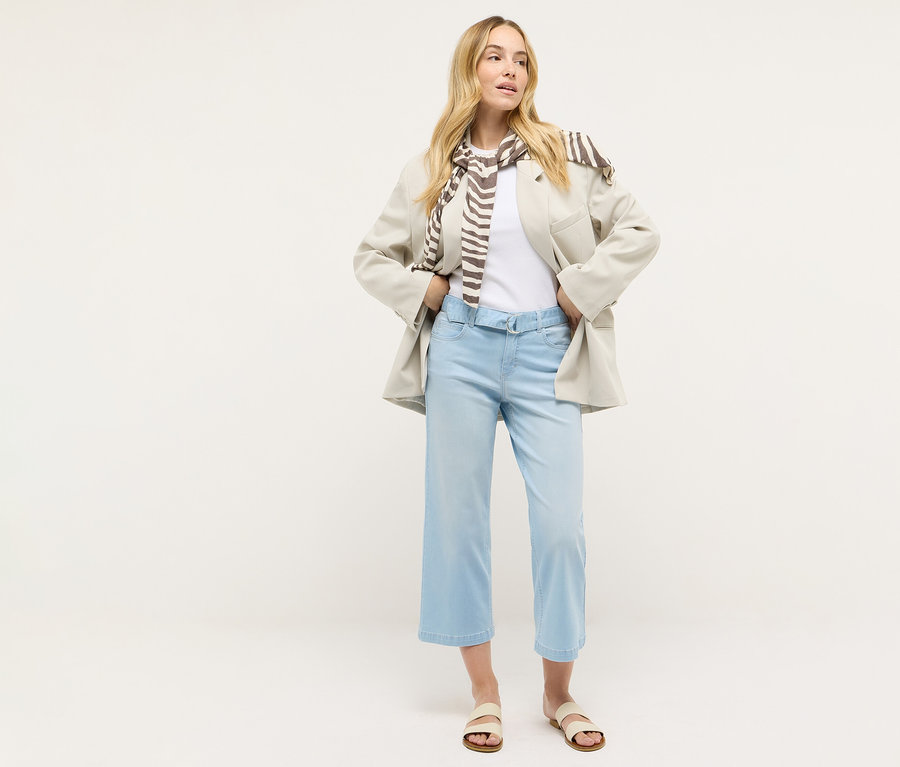 Une femme debout porte un jean Angels Denim « Linn City » cropped, bleached blue, un t-shirt blanc, un blazer et une écharpe.