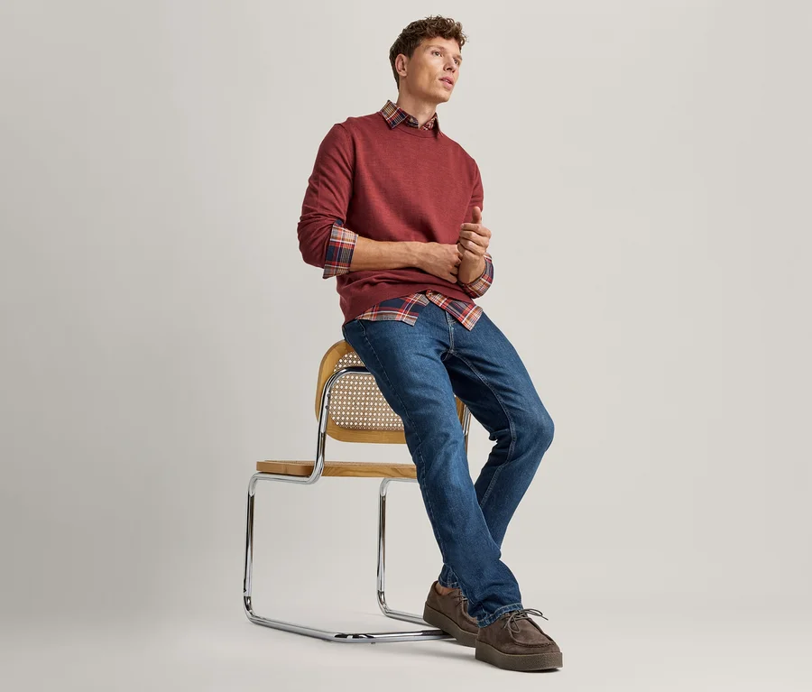 Jeune homme assis sur une chaise avec assise en bois tressé et cadre chromé, portant un pull rouge sur une chemise à carreaux, un jean bleu et des chaussures marron. Il regarde à droite.