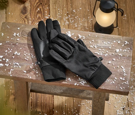Une paire de gants noirs avec des poignets côtelés et des paumes texturées repose sur un banc en bois rustique couvert de neige artificielle. Une lanterne allumée et des pommes de pin sont visibles sur le plancher en bois à l'arrière-plan.