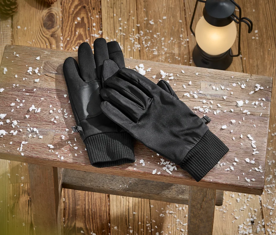 Une paire de gants noirs avec des poignets côtelés et des paumes texturées repose sur un banc en bois rustique couvert de neige artificielle. Une lanterne allumée et des pommes de pin sont visibles sur le plancher en bois à l'arrière-plan.