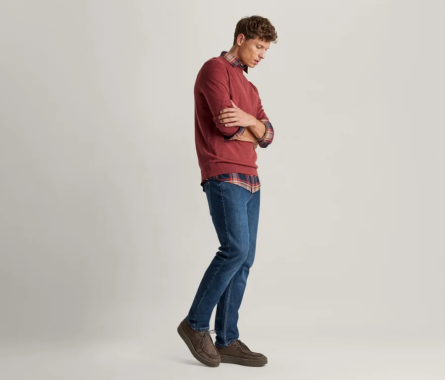 Un homme portant un pull rouge sur une chemise à carreaux, un jean bleu et des chaussures marron, debout les bras croisés et regardant vers le bas.