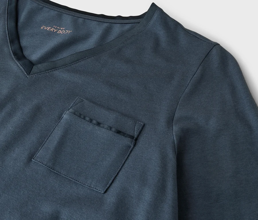 Gros plan sur un t-shirt gris-bleu foncé à col en V avec une poche poitrine. Le texte 'TCHIBO EVERY BODY' est visible à l'intérieur du col.