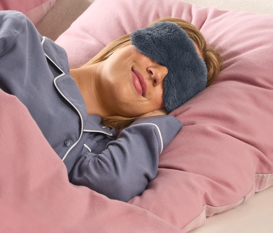 Une femme aux cheveux blonds dort paisiblement dans son lit, vêtue d'un haut de pyjama bleu avec un passepoil blanc et d'un masque de sommeil gris foncé et moelleux. Elle est couverte d'une couette rose et repose sa tête sur un oreiller rose.