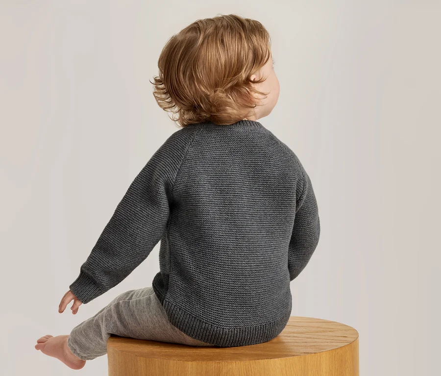 Bébé aux cheveux châtains clairs, vu de dos, assis sur un tabouret en bois. Le bébé porte un pull en tricot gris foncé et un pantalon gris clair.